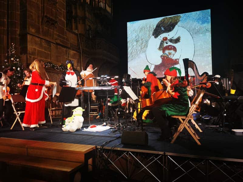 Orchestra in Christmas outfits with big screen in the background with the snowman hugging a boy on the screen