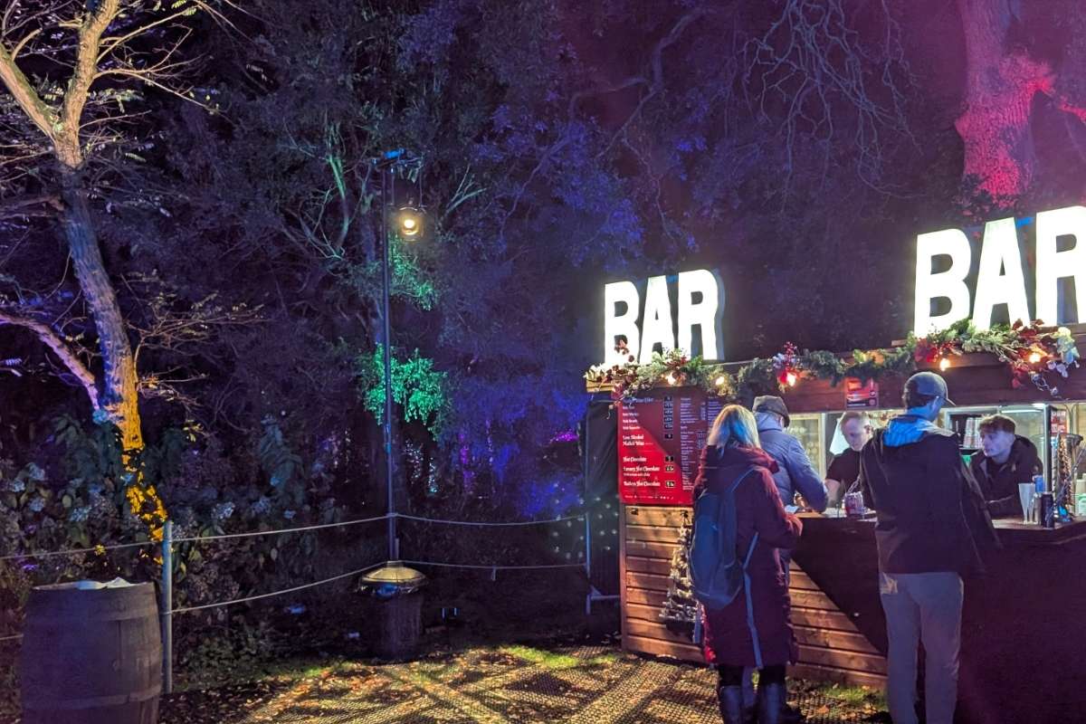A cozy outdoor bar kiosk is decorated with festive garlands and illuminated signs that read “BAR” in bold white letters. Visitors bundled in winter clothing chat with staff while ordering drinks, surrounded by trees bathed in colorful holiday lighting.