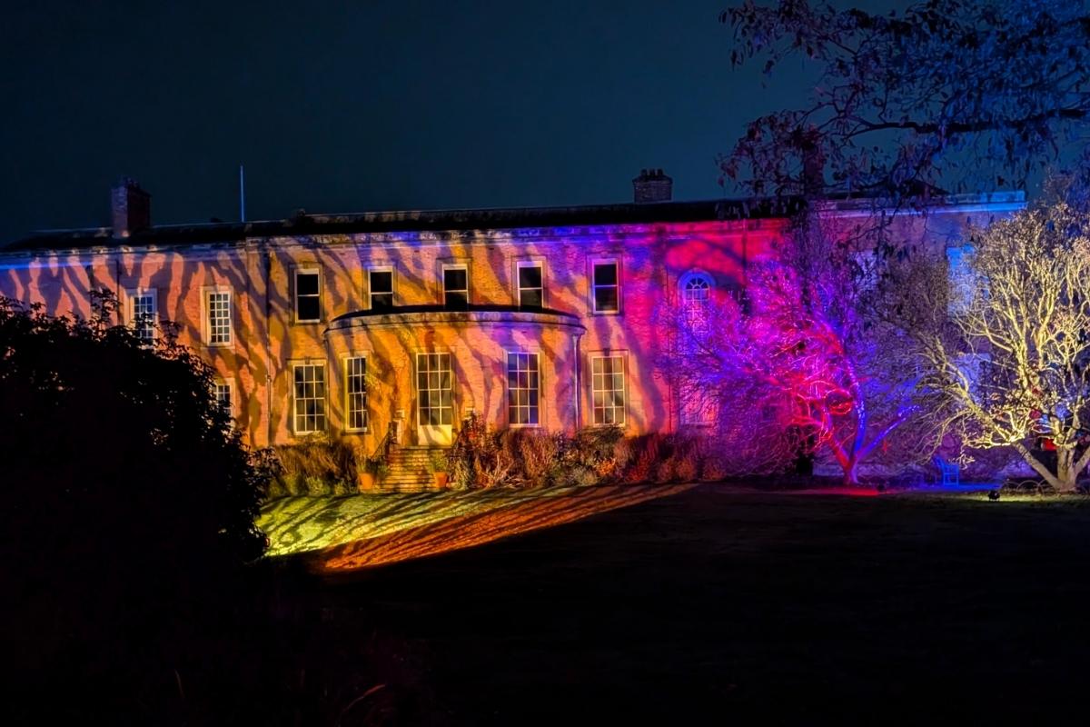 A stately brick mansion is illuminated at night with vivid projections of zebra-like orange, purple, and pink stripes across its facade. The surrounding trees are also lit in vibrant colors, creating a dramatic and festive atmosphere in the garden.