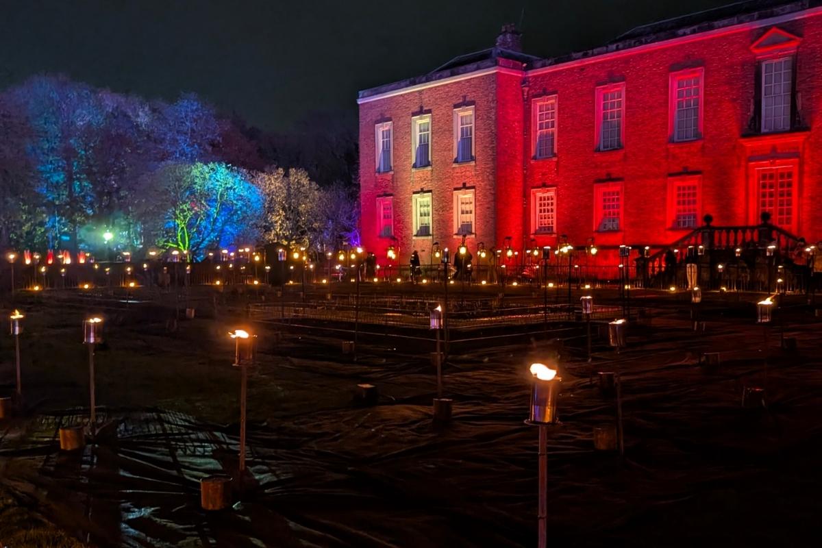 A historic brick building glows red at night, surrounded by a large outdoor display of torches flickering in the foreground. Trees in the background are lit in blue, green, and purple, adding a vibrant, atmospheric contrast to the warm firelight.