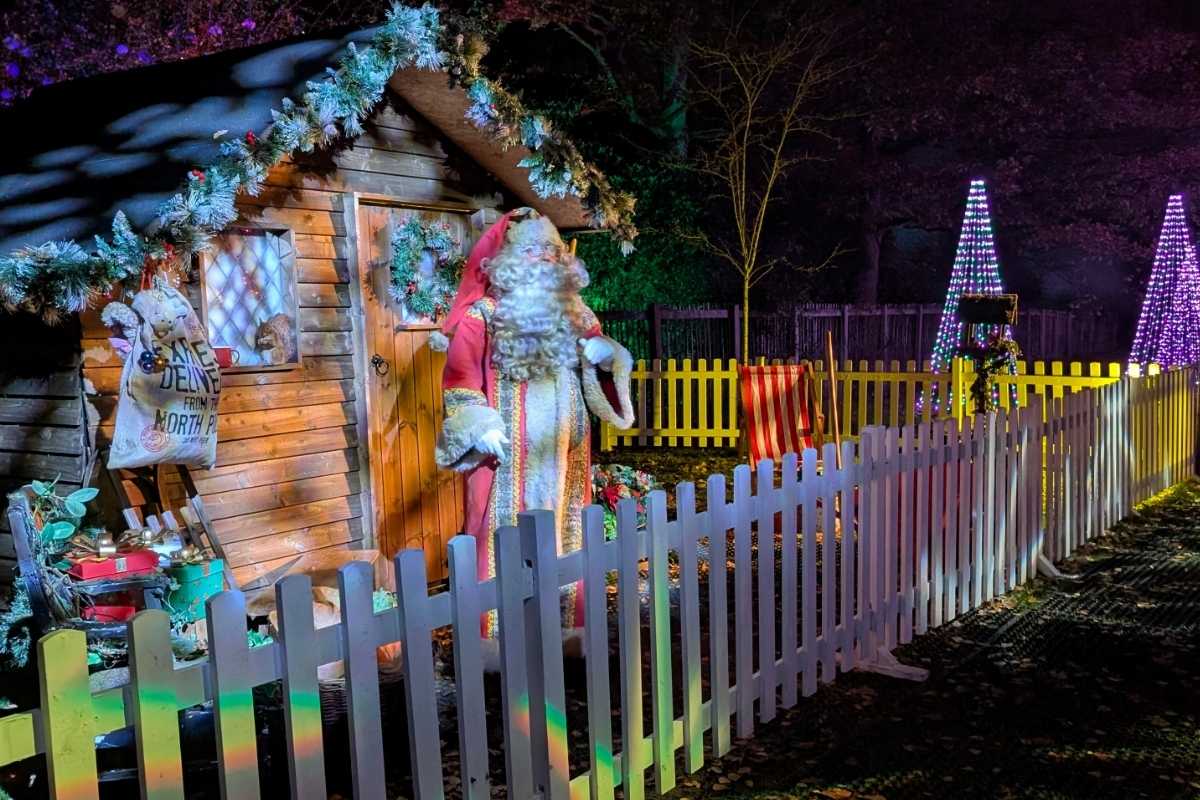 She Knows Alt Text said:A festive Santa Claus figure stands outside a wooden cabin decorated with garlands and a sack labeled “Special Delivery from the North Pole.” The scene is enclosed by a white picket fence, with glowing cone-shaped Christmas trees and colorful lights adding to the holiday charm in the background.