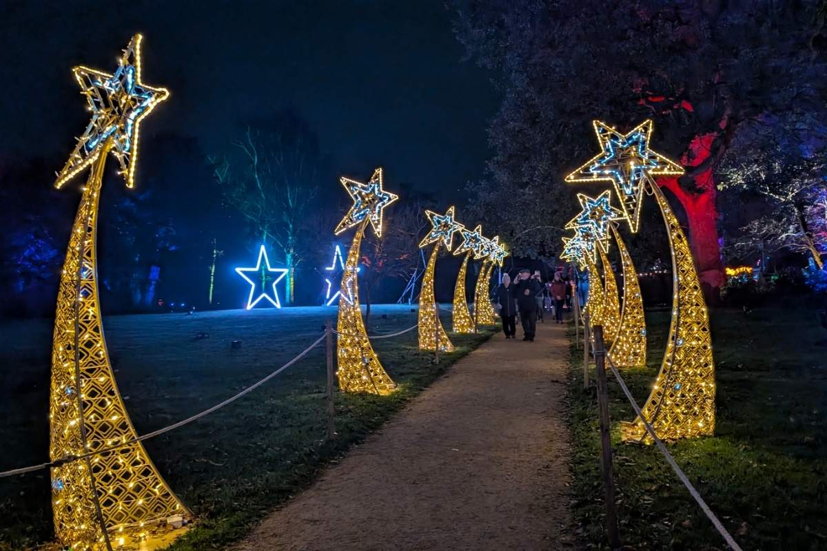 A nighttime walkway is lined with illuminated golden arches topped with glowing shooting stars, creating a festive tunnel effect. In the background, oversized neon stars shine in blue and white, while visitors stroll through the light display under a canopy of softly lit trees.