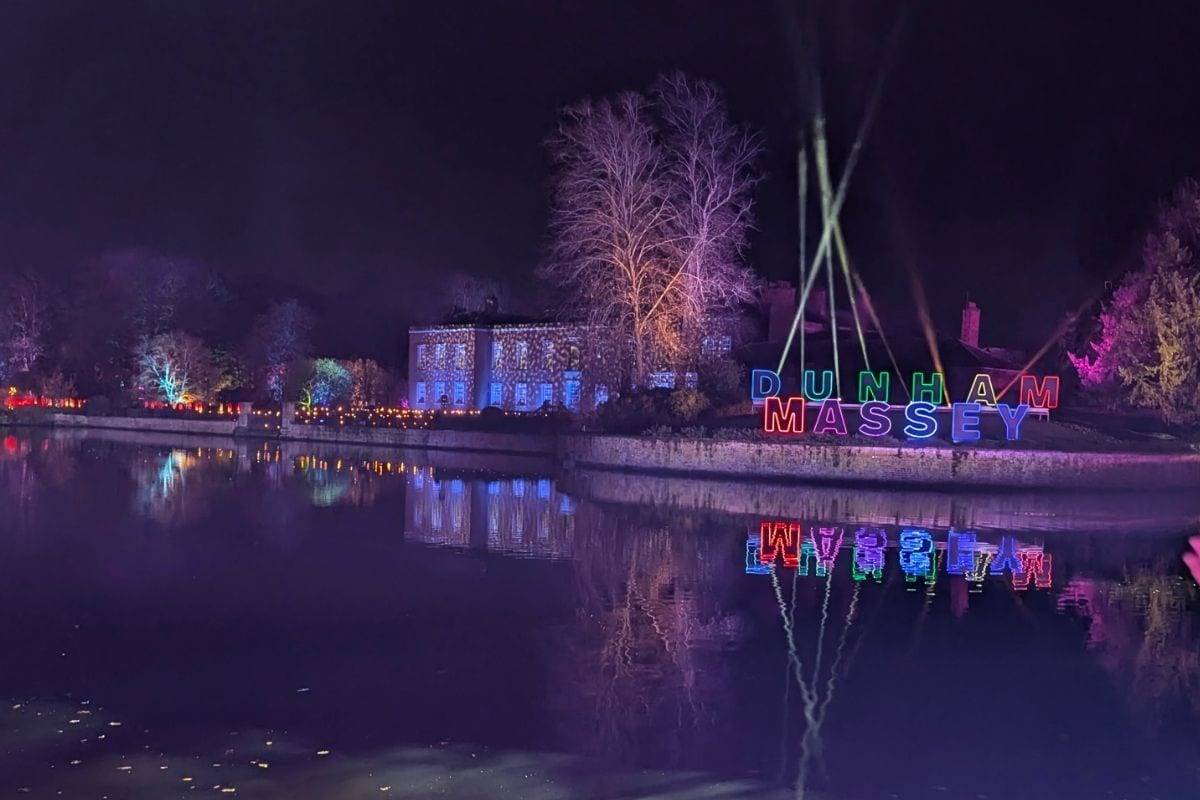 A nighttime view across a still body of water reflects vibrant lights and the illuminated sign "DUNHAM MASSEY" in bold, multicolored letters. Behind the sign, trees and a historic building are lit in purples and blues, creating a festive and magical scene.