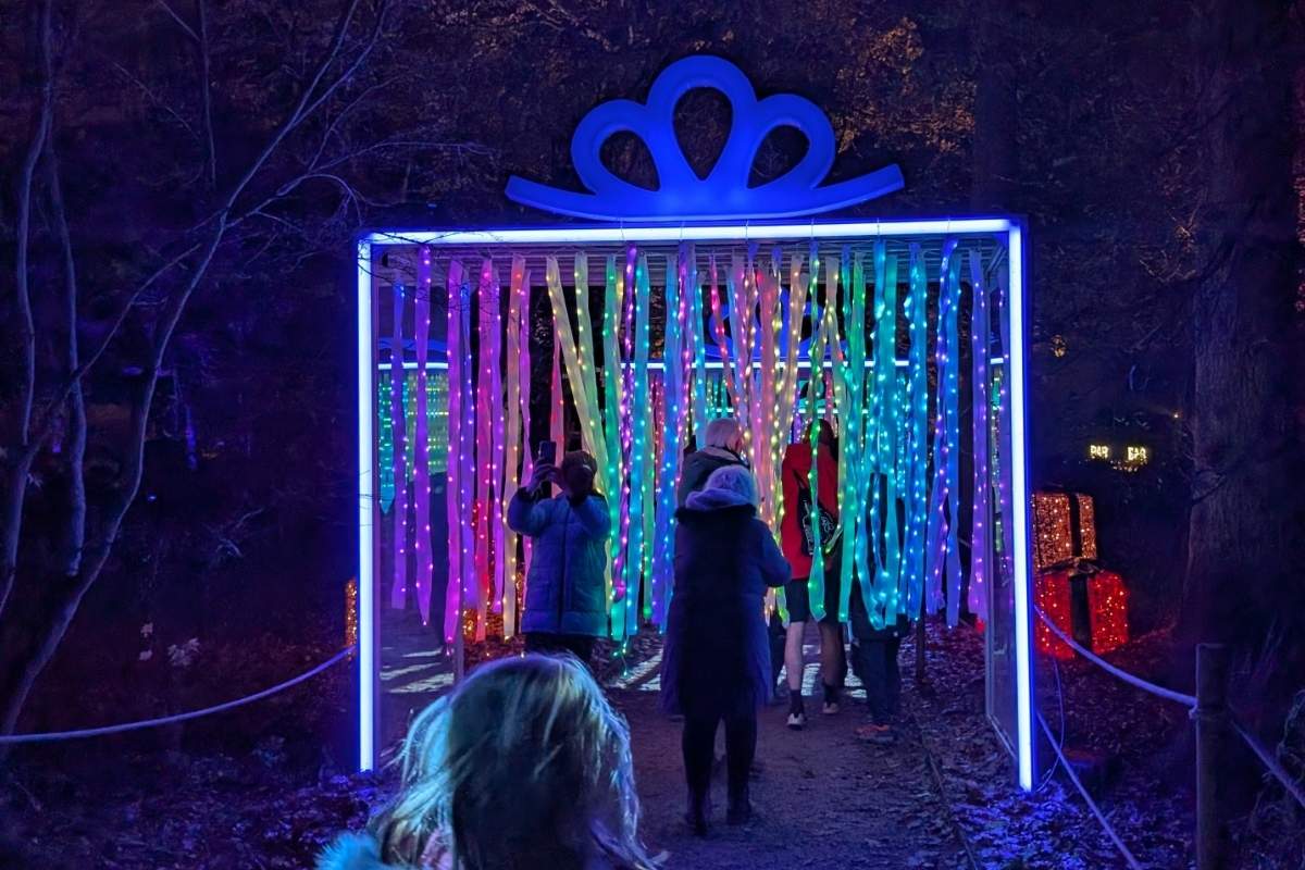 Visitors walk through a glowing tunnel shaped like a giant gift box, topped with a blue bow and filled with hanging LED ribbons in rainbow colors. The vibrant lights cast a festive glow on the path, with illuminated present decorations visible to the side.