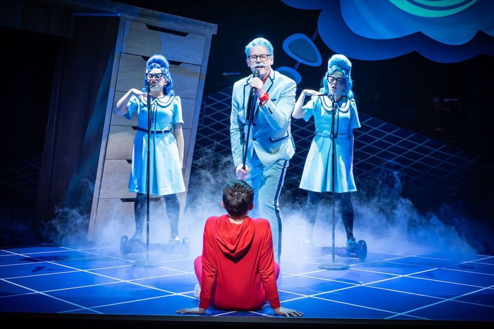 Boy dressed in red sat on floor with his back to us and a man and two women singing
