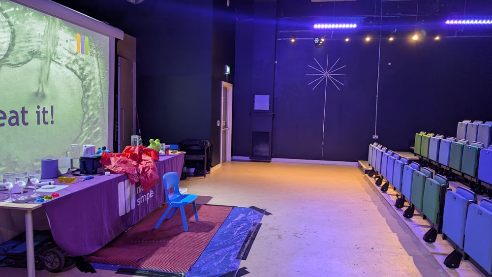 Theatre space with rows of seats at one side a screen and long table at the other side with lots of props