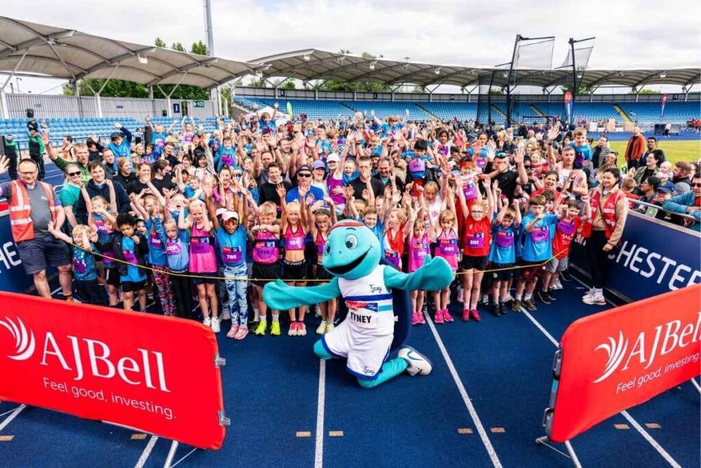 Large group of children and adults gathered on a blue running track at a stadium with their hands raised in the air. A turquoise mascot kneels at the front near red barriers that read "AJ Bell" and "Feel good investing."