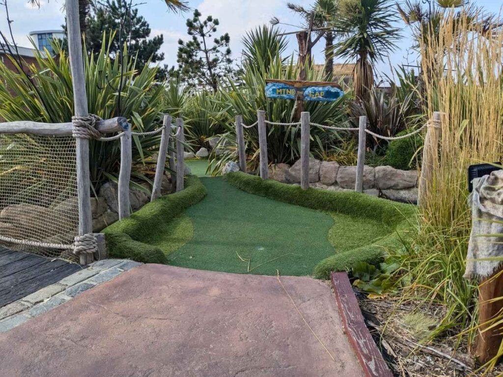 Crazy golf hole with lots of palms and grassess at either side and a sign that says Mind the gap