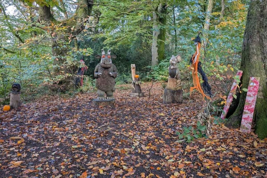 Forest clearing filled with autumn leaves and wooden sculptures of Gruffalo characters, including the Gruffalo, the mouse, and other figures on tree stumps. Halloween decorations like pumpkins, a witch costume, and painted planks are scattered around, adding a festive touch to the woodland setting.