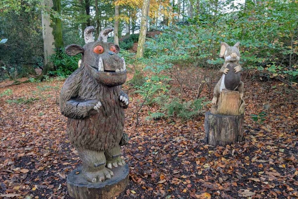 Wooden sculptures of the Gruffalo and the mouse from the children's book "The Gruffalo," set among autumn leaves on a forest trail. The Gruffalo stands on the left with wide orange eyes and tusks, while the mouse clutches a nut on a tree stump to the right.