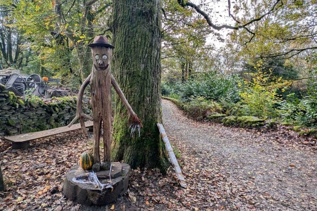 Wooden sculpture of Stick Man from the popular children’s book, wearing a brown hat and standing next to a large tree along a forest path. Autumn leaves cover the ground, with a gourd and fake spider webs placed at the statue’s base as seasonal decorations.
