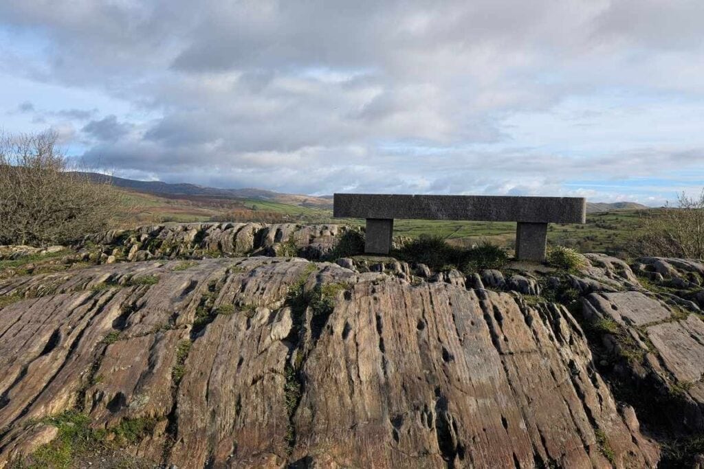 Stone bench perched atop a rugged, striated rock outcrop at Orrest Head, overlooking rolling green hills under a partly cloudy sky. The bench offers a quiet spot to rest and take in the expansive countryside views.