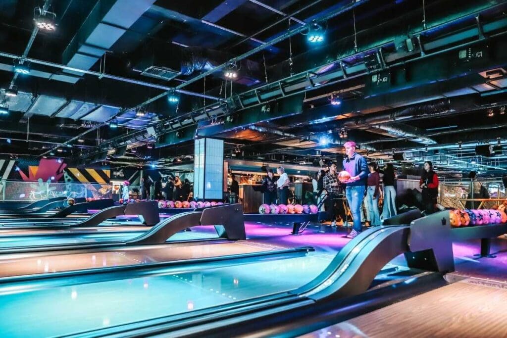 A modern bowling alley with people preparing to bowl, glowing blue lanes, and racks of orange bowling balls, all under dramatic ceiling lights and colorful decor.
