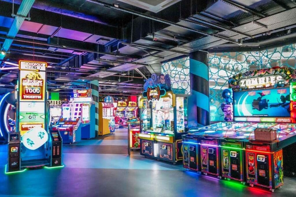 A wide view of vibrant arcade cabinets like "Monopoly," "Zombies," and claw machines, set in a glowing, futuristic game zone with patterned pillars and multicolored lighting.