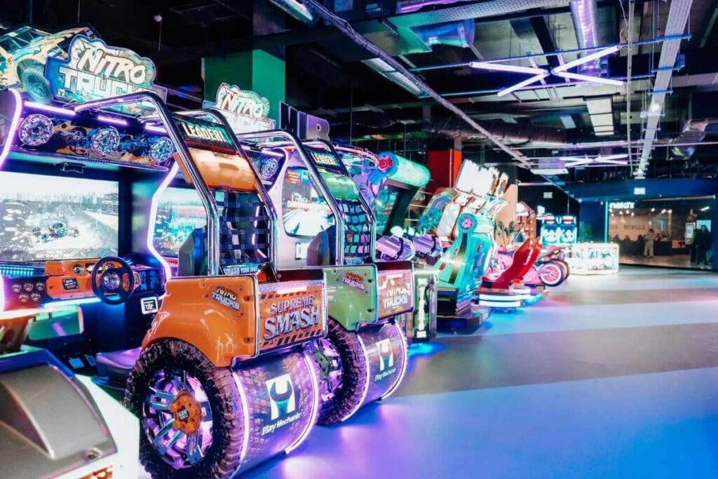 Rows of brightly lit racing arcade machines, including "Nitro Trucks" and "Supreme Smash," with oversized truck-themed seats and neon lights glowing under dark industrial ceilings.