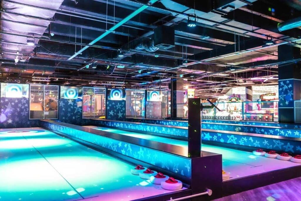 A glowing curling game setup with two long, neon-lit lanes surrounded by star-patterned panels, curling-style pucks lined up at the ends, and arcade games visible in the background.