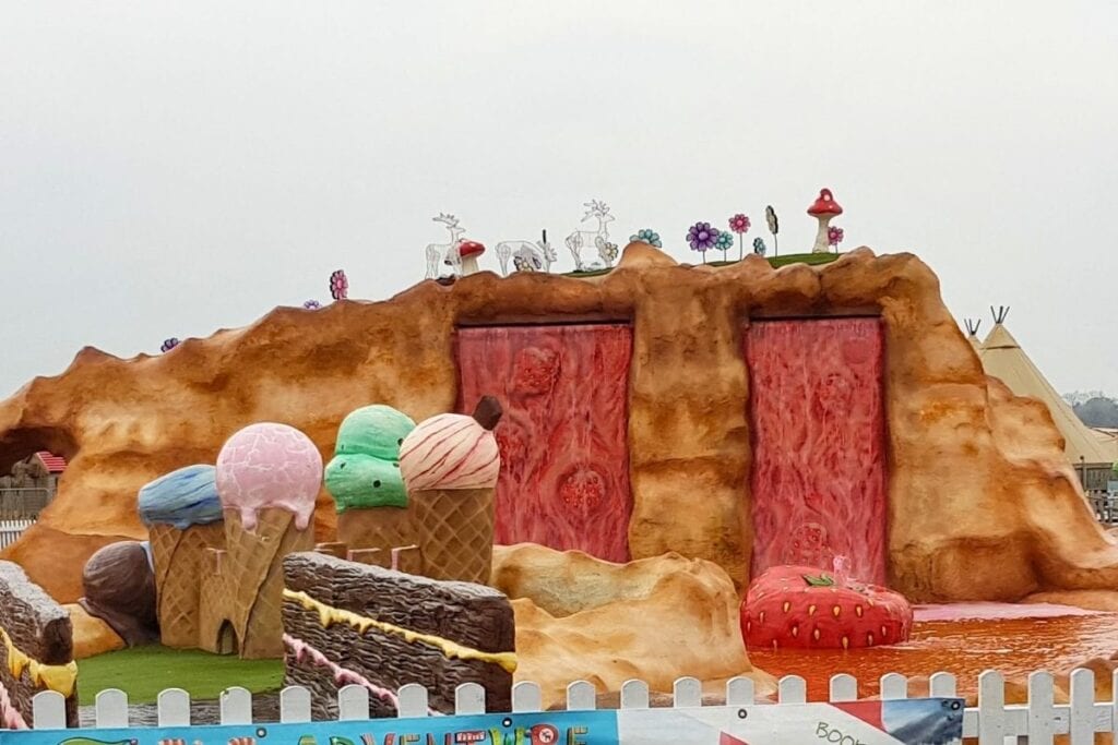 Giant ice cream cone sculptures in pastel blue, pink, green and vanilla colors stand in front of a fantasy themed rock formation at The Ice Cream Farm. Pink waterfall features flow down the center of the display, with oversized strawberries, mushrooms and colorful flowers decorating the scene behind a white picket fence.