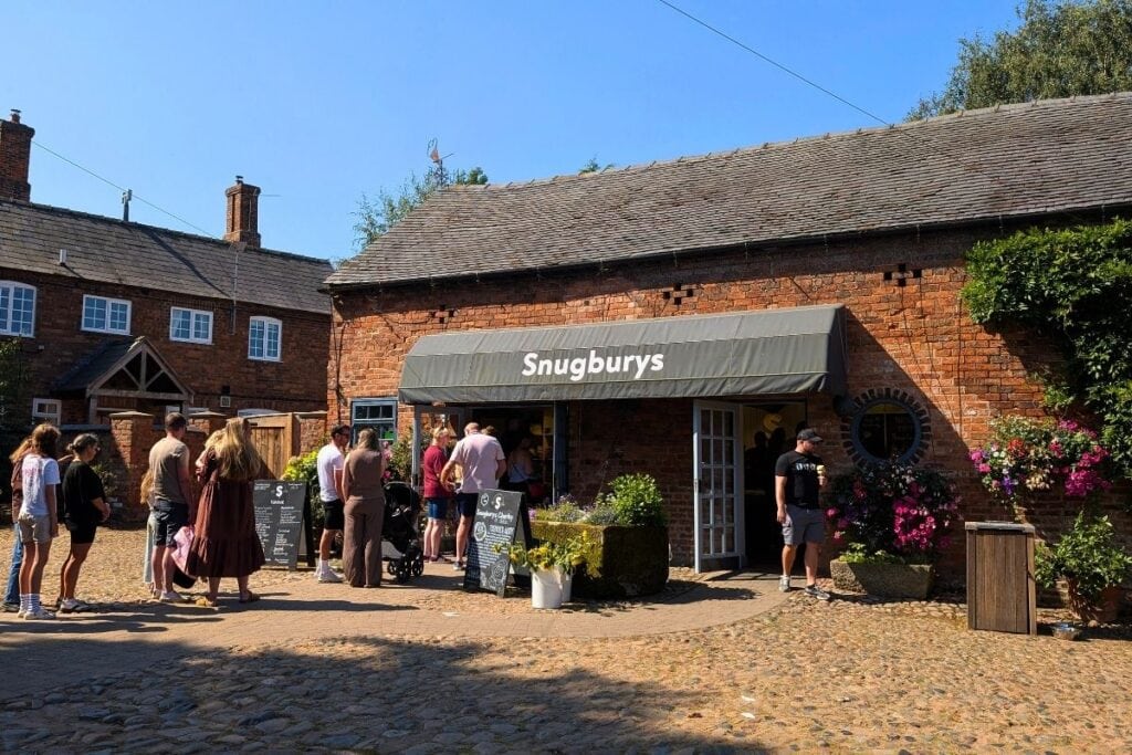 Visitors queue outside Snugburys on a sunny day, gathered in front of a red brick farm building with a grey awning displaying the shop name. Flower displays and chalkboard signs line the entrance as people wait to buy ice cream in the cobbled courtyard.