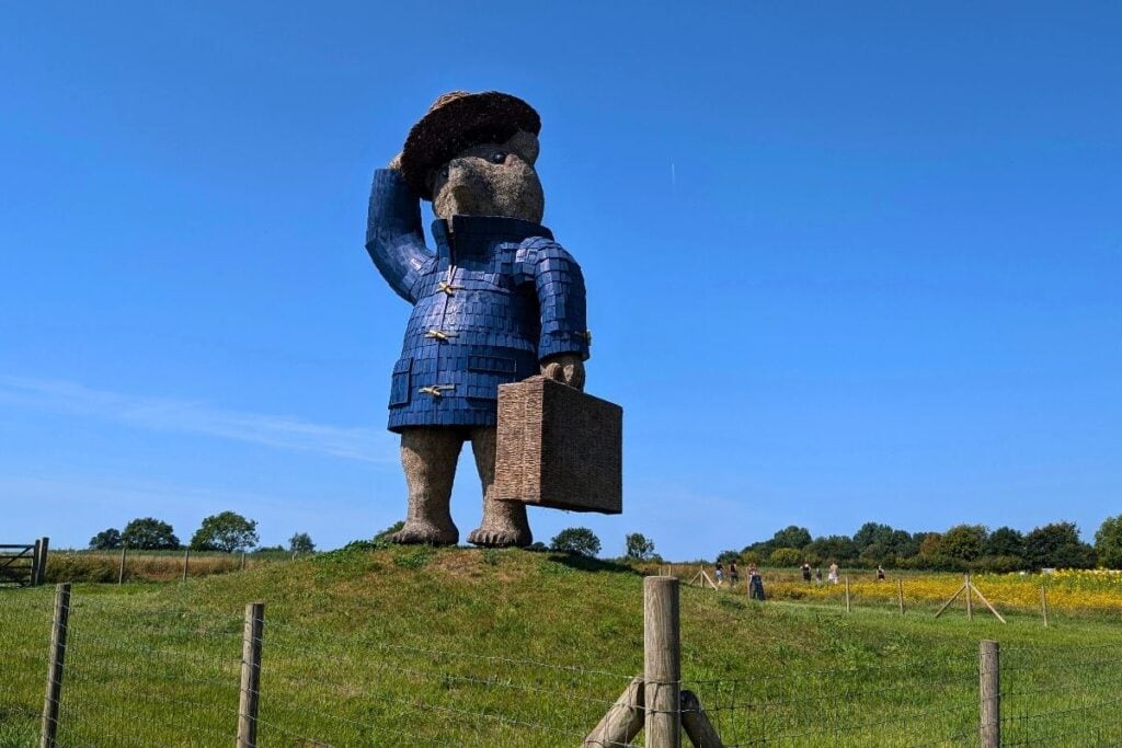 Giant Paddington Bear sculpture stands on a grassy hill under a clear blue sky, wearing his signature blue coat and red hat while holding a suitcase. Visitors walk through the surrounding fields in the distance, highlighting the scale of the landmark at Snugburys.