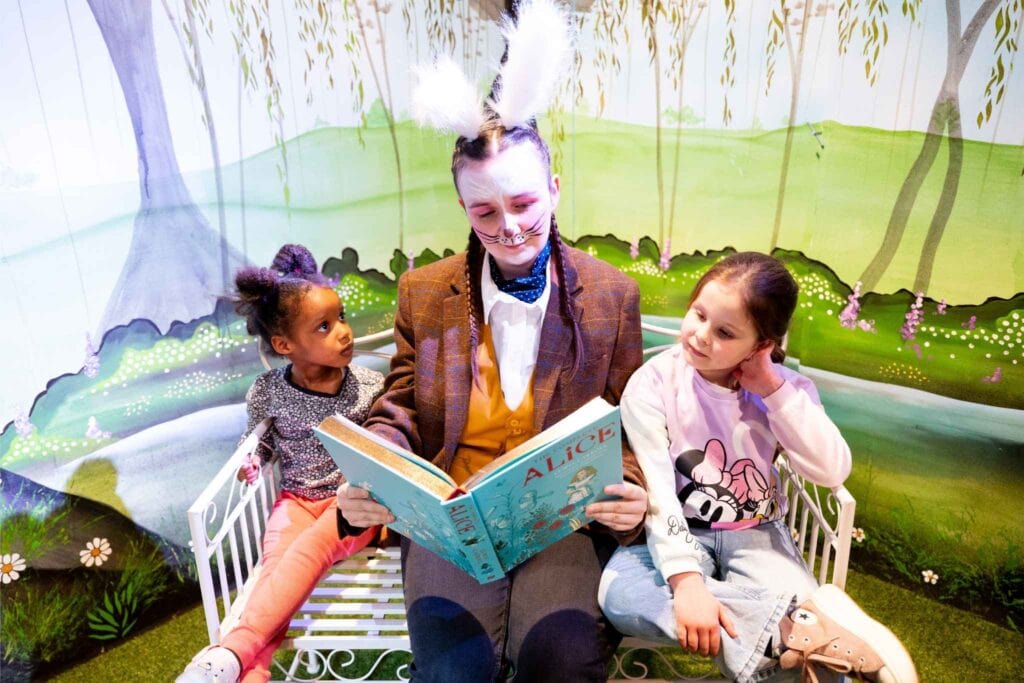 Lady dressed as a rabbit sat on bench reading an oversized book with 2 young girls