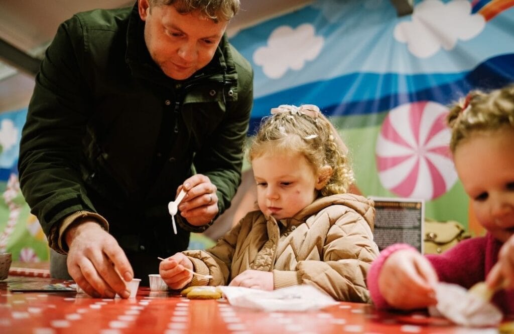 Child decorating a biscuit with icing at Walby Farm Park Christmas crafts activity, helped by an adult, with a festive candy-themed backdrop.