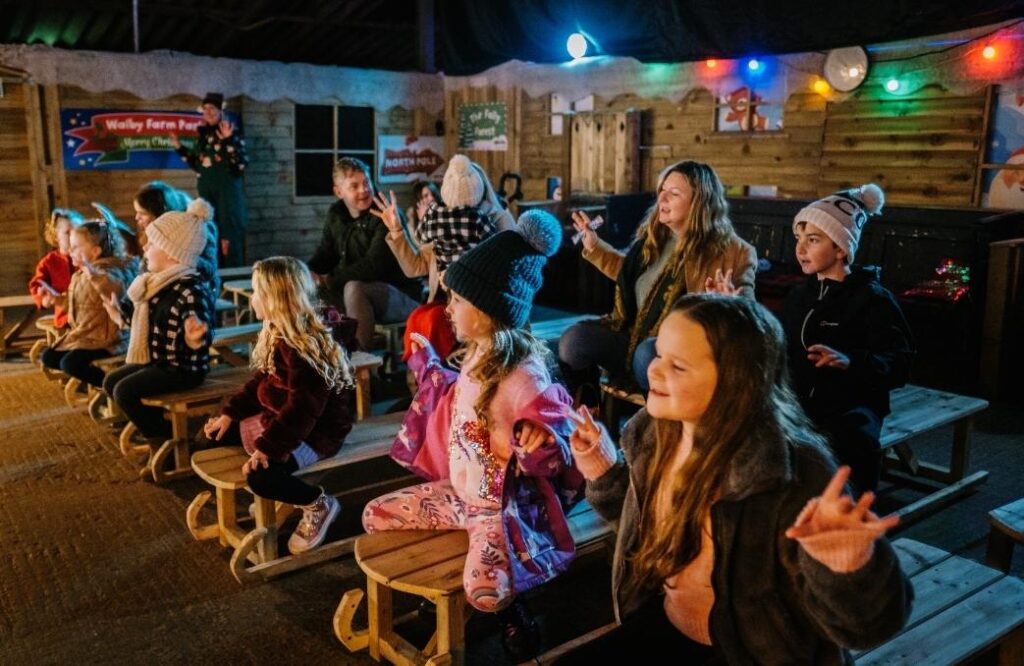 Families and children sitting on benches at Walby Farm Park, enjoying a lively Christmas show with festive decorations and colourful lights.